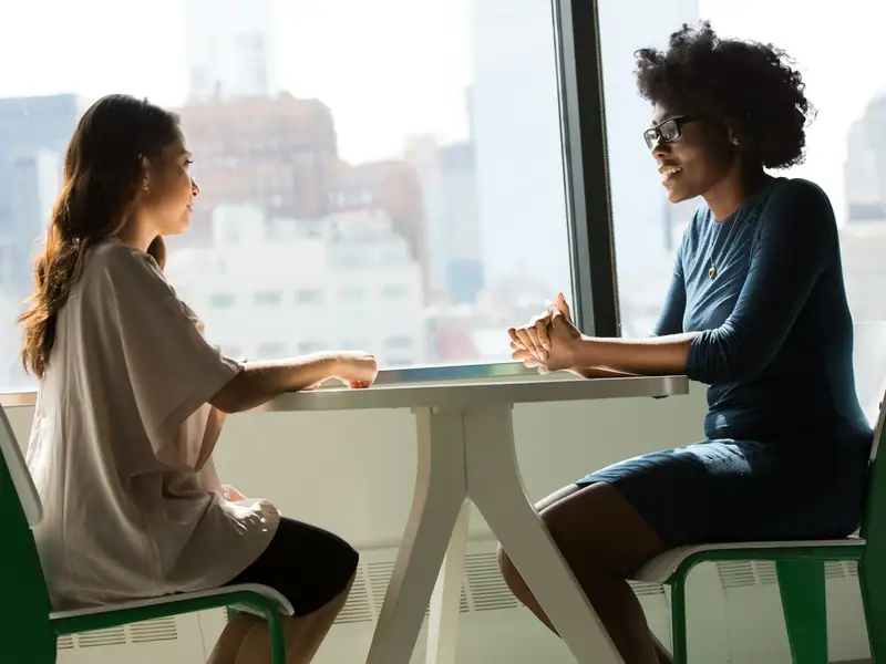 Two colleagues in conversation at a table by a bright office window